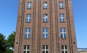 Sechsstöckiges Backsteingebäude mit weißen Sprossenfenstern und dekorativem Ziegelmuster im oberen Bereich, blauer Himmel.
