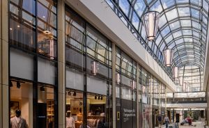 Glasüberdachte Einkaufspassage mit Geschäften, Pendelleuchten, Sitzbank und Besuchern bei Tageslicht.