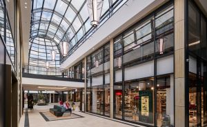 Helles Einkaufszentrum mit Glasdach, eleganten Hängelampen, zwei Etagen, Geschäften hinter Glasfronten und Sitzmöbeln im Gang.