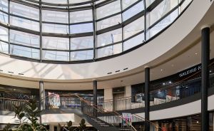 Rundes Einkaufszentrum-Atrium mit Glasdach, goldenes Oldtimer-Exponat, Treppenaufgang, mehrere Etagen, Pflanzen.