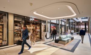 Einkaufszentrum mit Besuchern, modernem Interieur, Deckenbeleuchtung, Geschäften und einer mintfarbenen Sitzbank im Mittelgang.