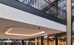 Modernes Einkaufszentrum mit Glasdach, Obergeschoss-Galerie, grüner Sitzbank und zwei Personen vor Ladengeschäften.