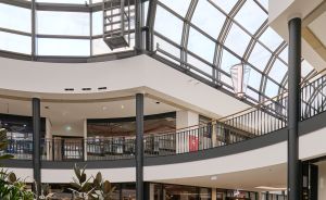 Modernes Einkaufszentrum mit Glasdach, zwei Etagen, Rundgalerie und drei Personen im Erdgeschoss.