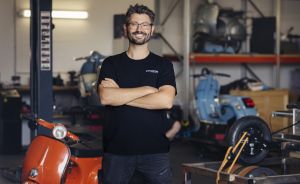 Mann mit Brille und schwarzem T-Shirt steht mit verschränkten Armen in einer Werkstatt mit Motorrollern.