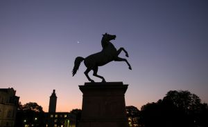 Silhouette einer Pferdestatue auf Sockel im Abendhimmel, dahinter Gebäude und Bäume, Mondsichel sichtbar.