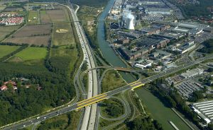 Luftaufnahme einer Autobahnkreuzung mit gelber Brücke über einen Kanal, neben Industriegebiet mit Schornsteinen.