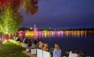 Menschen sitzen abends am beleuchteten Seeufer, im Hintergrund bunte Lichtreflexe auf dem Wasser und ein Springbrunnen.