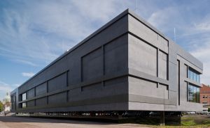 Modernes Gebäude aus dunklem Beton, auf Stützen erhöht, mit geometrischer Fassade, unter blauem Himmel mit Wolken.