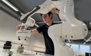 „Junger Mann in schwarzem Shirt justiert den Arm eines weißen Industrieroboters auf einem Arbeitstisch."