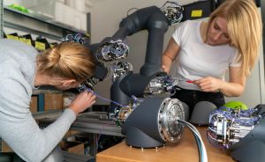 Zwei Frauen arbeiten an einem schwarzen Roboterarm mit Kabeln auf einem Tisch in einem Lagerraum.