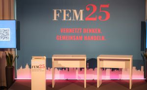 Bühnenaufbau mit blauem Hintergrund, Aufschrift „FEM25 – Vernetzt denken. Gemeinsam handeln." und drei weißen Tischen.
