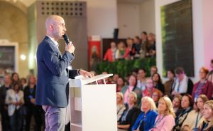 Mann im Blazer spricht an weißem Rednerpult mit Mikrofon vor großem Publikum in einem Saal.