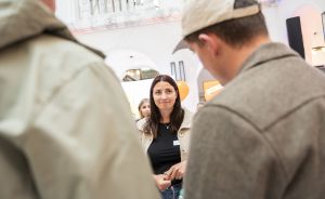 Frau mit schwarzem Shirt und Namensschild spricht mit zwei Personen in beigen Jacken in einem hellen Gebäude.