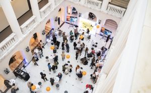 Luftaufnahme einer Veranstaltung in einem weißen Atrium mit Säulen, Stehtischen, orangen Ballons und etwa 50 Personen.