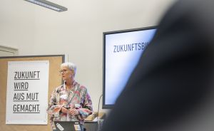 Frau mit kurzen grauen Haaren präsentiert vor Leinwand mit „Zukunftsbild", Plakat „Zukunft wird aus Mut gemacht." im Hintergrund.