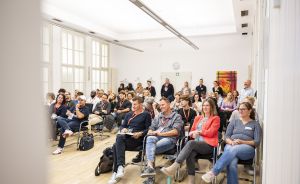 Gruppe von Personen sitzt und steht in einem hellen Raum bei einer Veranstaltung, viele tragen Namensschilder.