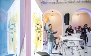 Person mit Mikrofon spricht vor Publikum in hellem Veranstaltungsraum mit Stellwänden und orangem Ballon.