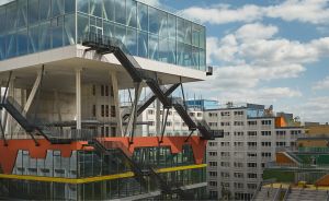 Modernes Gebäude mit Glasaufbau auf Stahlstützen, orangefarbenen Elementen und Außentreppen, dahinter mehrstöckige Bürogebäude.