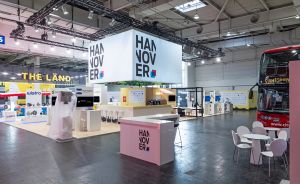 Messestand „Hannover" in einer Halle mit rosa Tresen, weißem Kubus, Sightseeing-Bus rechts und Firmenständen im Hintergrund.