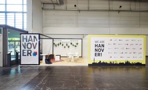 Messestand mit „HANNOVER"-Schriftzug und gelbem „We Are Hannover!"-Banner mit Stadtsilhouette in einer Messehalle.