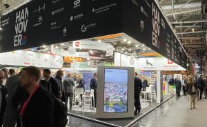 Messestand Hannover mit schwarzem Dach und Logos in großer Halle, Besucher laufen vorbei, Bildschirm im Vordergrund.
