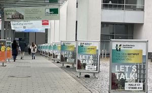 Messehalle mit Werbebannern für Techfactory Hannover (Halle C1, Stand 410) und AL|HO. Fußgänger auf gepflastertem Weg.