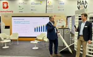 Zwei Männer stehen auf einer Messe vor einer Leinwand mit Balkendiagramm, einer hält ein Windrad-Modell.
