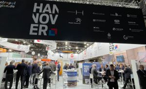 Messestand Hannover mit großem Schriftzug, Besucher in Halle, Firmenlogos auf Banner, modernes Hallendach.
