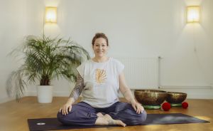 Frau sitzt im Schneidersitz auf Yogamatte, lächelt, trägt weißes T-Shirt, hat Tattoos, Klangschalen im Hintergrund.