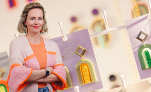 Frau mit verschränkten Armen vor einem Display mit bunten Ohrringen aus Acryl und Gold auf lila Karten.
