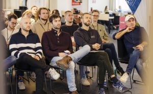 Gruppe junger Menschen sitzt auf Stühlen in einem Büro und hört einer Präsentation zu. Im Hintergrund ein Banner.