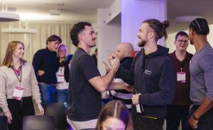 Zwei Männer begrüßen sich lächelnd per Handschlag in einem Raum mit mehreren Personen im Hintergrund, die Namensschilder tragen.