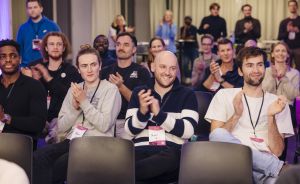 Publikum applaudiert bei einer Veranstaltung; Männer und Frauen sitzen und stehen in Reihen, einige tragen Namensschilder.
