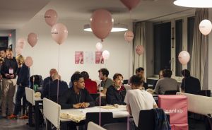 Personen sitzen an Tischen in einem Raum mit rosa Ballons, auf denen "leanlab" steht. Poster hängen an der Wand.
