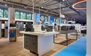 Modernes Ladengeschäft mit weißen Verkaufstresen, Monitoren, Kaffeemaschinen und Holzboden unter orangefarbener Ringlampe.