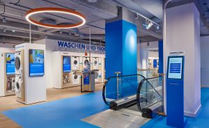 Elektronikabteilung mit Waschmaschinen, blauem Boden, Rolltreppe, Digitalanzeigen und Schild „Waschen und Trocknen".