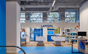 Elektronikgeschäft mit blauem Bodenbelag, Laptops-Schild, Produktdisplays auf weißen Tischen und Wandregalen mit Zubehör.
