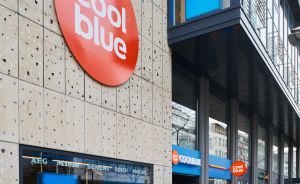 Coolblue-Filiale mit rotem Markenlogo an Steinfassade, Schaufenster mit blauem Display, eine Person betritt das Geschäft.