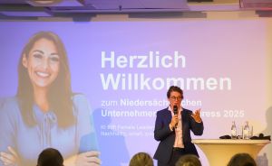 Person spricht mit Mikrofon vor Publikum. Präsentation zeigt „Herzlich Willkommen zum Niedersächsischen Unternehmerkongress 2025".