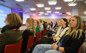 Frauen sitzen in einem Konferenzraum mit roten Stühlen und schauen auf eine Präsentationsleinwand.