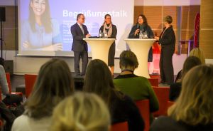 Podiumsdiskussion mit vier Personen an Stehtischen vor Publikum in rotem Konferenzsaal, Präsentationsfolie im Hintergrund.