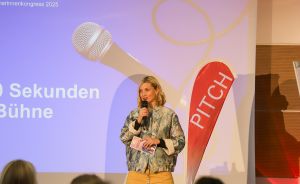 Frau spricht mit Mikrofon vor Publikum, Leinwand zeigt „90 Sekunden Bühne", rotes PITCH-Banner im Hintergrund.