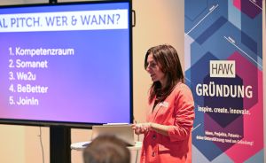Frau in rotem Blazer präsentiert vor Leinwand mit Pitchliste. Banner mit "HAWK Gründung – Inspire. create. innovate."