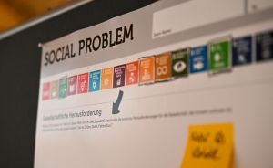 Whiteboard mit der Aufschrift „Social Problem", UN-Nachhaltigkeitsziele-Icons und deutschen Texten, gelbes Haftnotiz-Zettel.