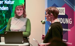 Zwei Frauen auf einer Bühne: eine spricht ins Mikrofon, eine steht am Laptop. Banner mit „Gründung" im Hintergrund.