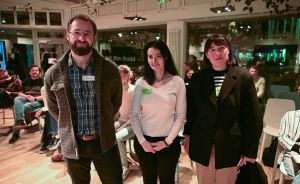 Drei Personen mit Namensschildern stehen nebeneinander in einem belebten Veranstaltungsraum mit grüner Beleuchtung.