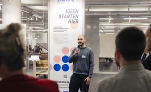 Mann mit Mikrofon spricht vor Publikum vor einem "Starting Business – Ideen starten hier"-Banner in einem Innenraum.