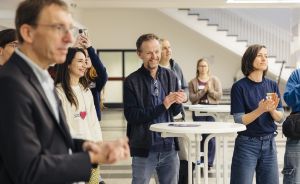 Mehrere Personen stehen in einem hellen Raum und applaudieren; eine Frau fotografiert mit dem Smartphone.