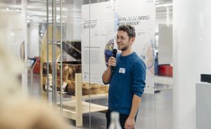 Mann mit blauem Pullover und Brille spricht in Mikrofon vor Ausstellungstafeln in einem Museum.