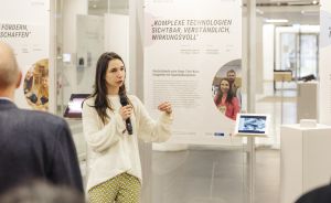 Frau mit Mikrofon spricht vor Publikum in Ausstellungsraum, hinter ihr deutschsprachige Infotafel über komplexe Technologien.
